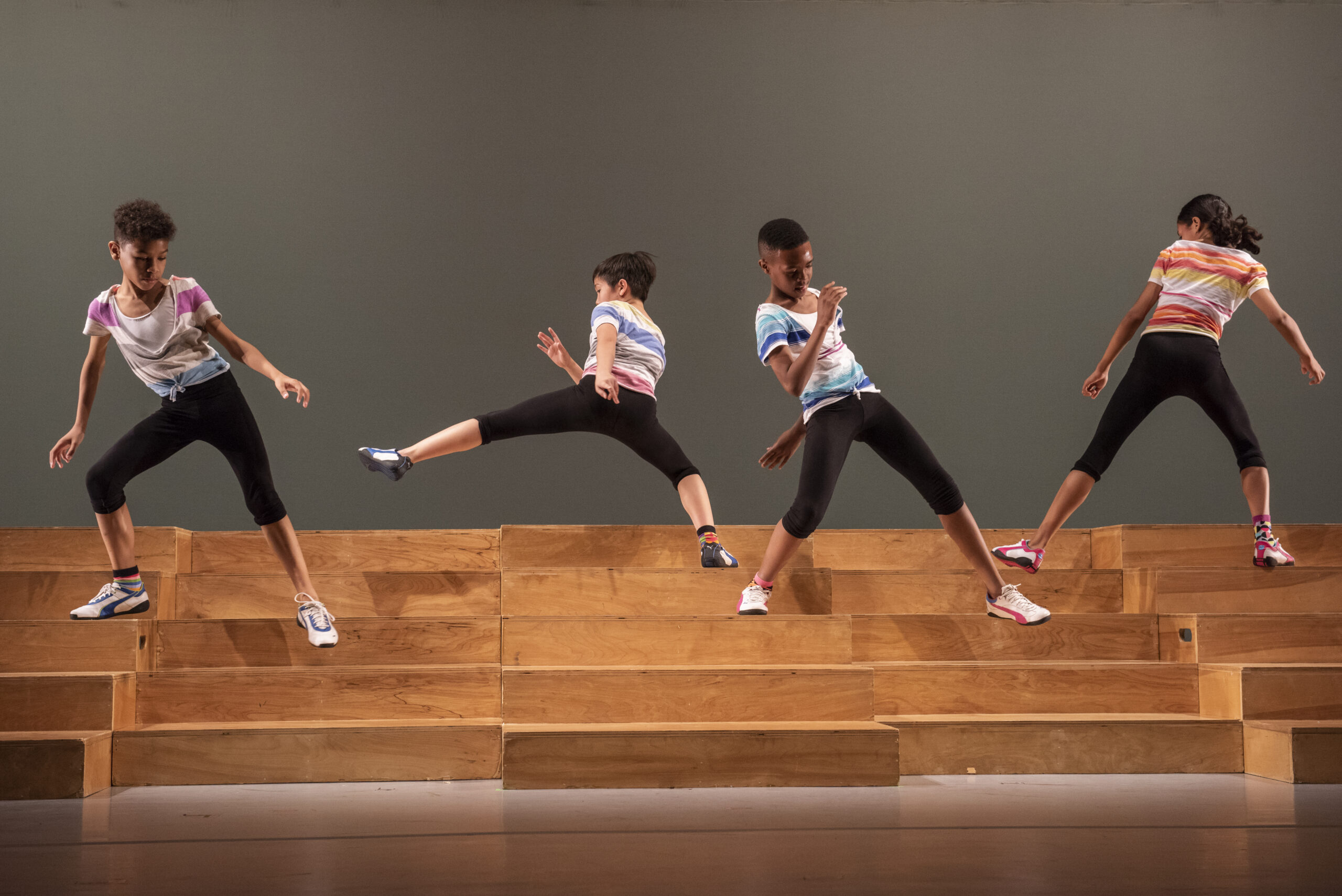 Four young dancers wearing colorful t-shirts and black leggings perform dynamic moves on a wooden staircase. The dancers are positioned in varied energetic stances, showcasing synchronized choreography against a simple backdrop.